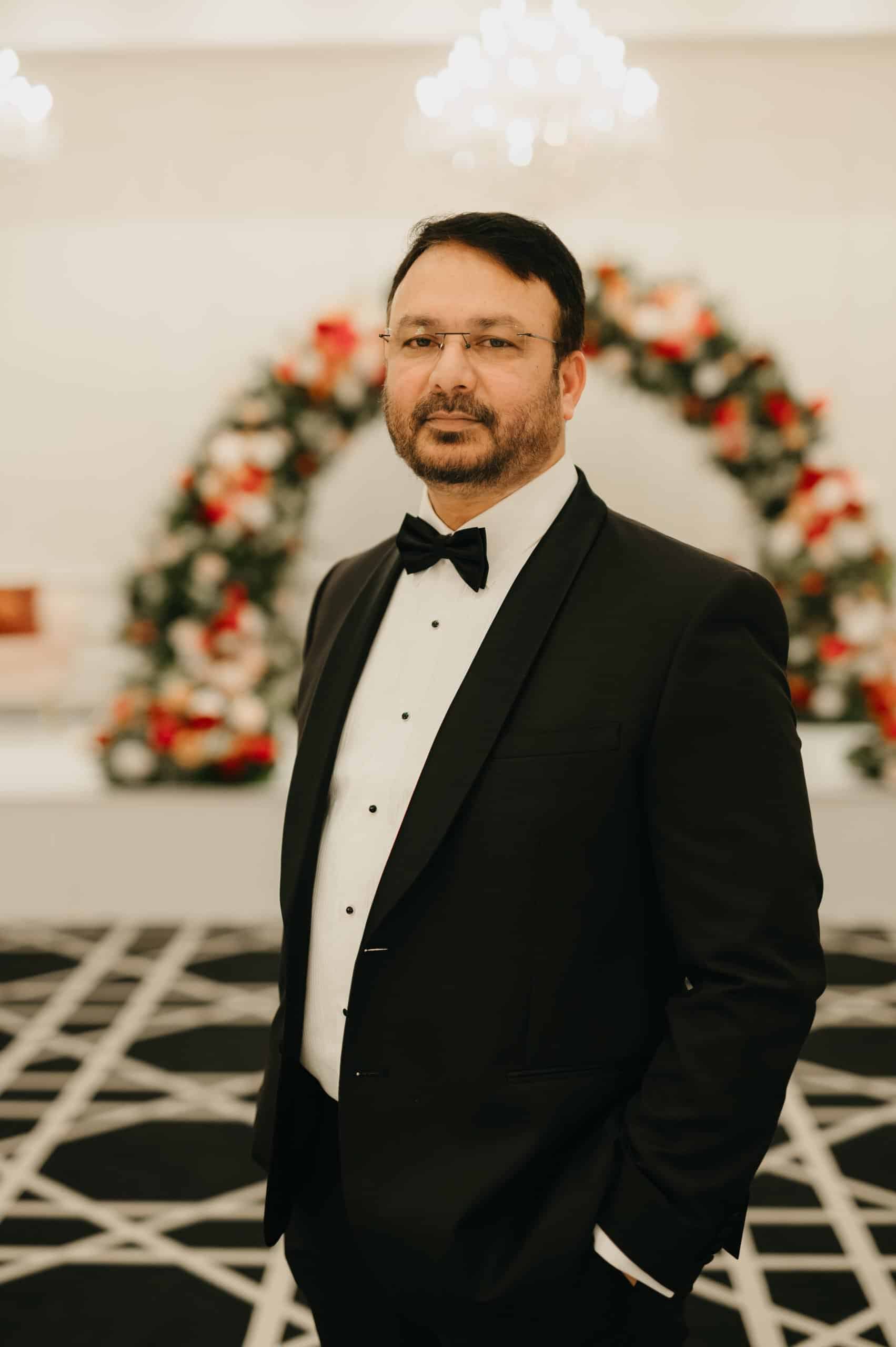 Confident man in black tuxedo with bow tie at wedding venue, standing in front of floral arch, representing professionalism and style.